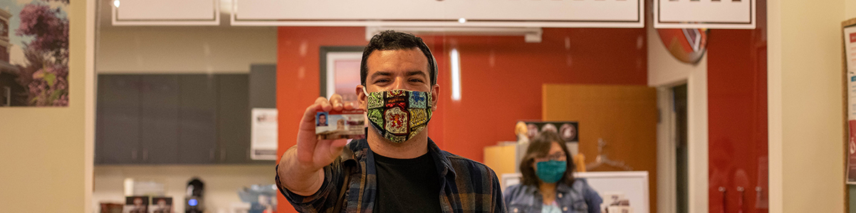 Student wearing face mask holds his CougarCard towards the camera. He is standing in front of the CougarCard Center.